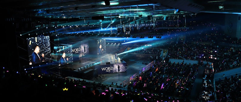 A large indoor arena filled with an audience watching a live performance. The stage features large screens displaying a performer and graphics, with bright spotlights and ambient lighting creating a dynamic atmosphere. The crowd is holding glow sticks, adding vibrant colors to the scene.
