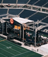 A large outdoor stage is set up inside a stadium, equipped with tall scaffolding, lights, and multiple large display screens. The seats in the stadium are mostly empty. The field is marked with numbers and lines typical of a sports venue. A few people are visible near the stage and on the field.