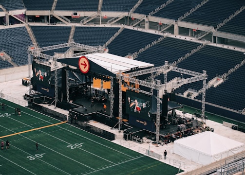 A large outdoor stage is set up inside a stadium, equipped with tall scaffolding, lights, and multiple large display screens. The seats in the stadium are mostly empty. The field is marked with numbers and lines typical of a sports venue. A few people are visible near the stage and on the field.