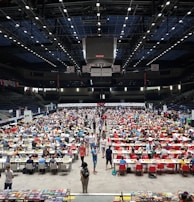 An indoor sports arena filled with rows of tables, each occupied by people engaged in competitive activities. The ceiling is high, with numerous lights illuminating the large space. The audience seating on either side is empty, and there are flags hanging from the upper level.