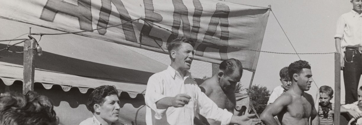 A group of men, some shirtless, are gathered around a platform or stage. A man in a white shirt appears to be speaking or gesturing to a crowd. A large sign with the word 'ARENA' is prominently displayed in the background. The scene seems to be set outdoors on a sunny day.