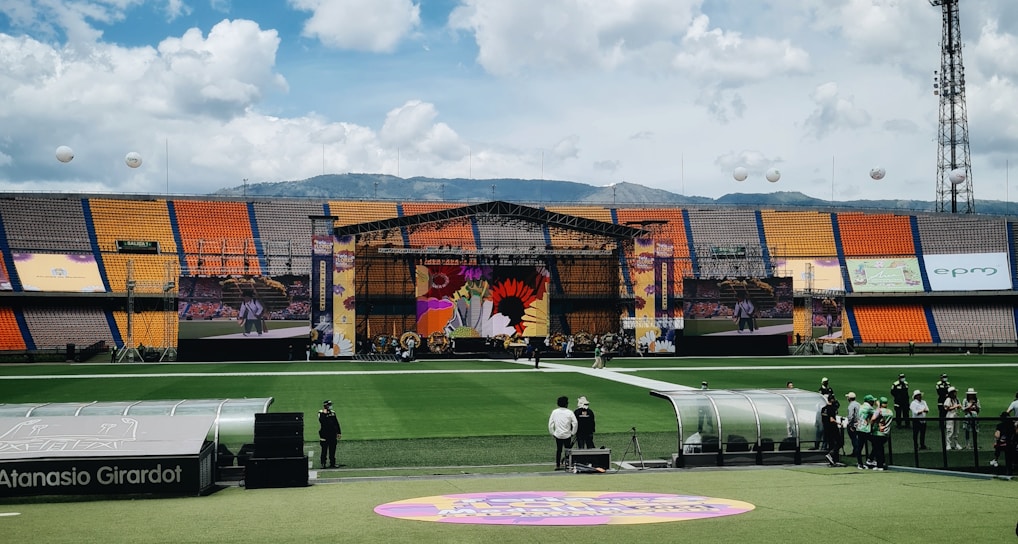 A sports stadium with a large stage set up on the field, hosting an outdoor event. Spectators wearing colorful hats sit in the foreground. The seating areas display vibrant colors, and a clear blue sky with scattered clouds is visible. Large screens display graphics related to the event.