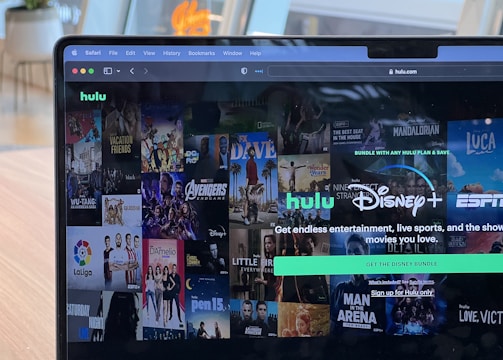 A laptop screen displays a web page featuring a mix of movie and TV show covers, highlighting popular titles and a Hulu and Disney+ bundle promotion. There are visible icons and images of films and shows alongside promotional text offering entertainment and live sports.