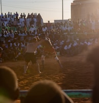 A vibrant wrestling match taking place outdoors, surrounded by a large crowd of spectators. The atmosphere is charged with excitement as people stand and sit around the dirt arena, intently watching the competitors. The sun is setting, casting a warm glow over the scene.