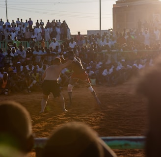 A vibrant wrestling match taking place outdoors, surrounded by a large crowd of spectators. The atmosphere is charged with excitement as people stand and sit around the dirt arena, intently watching the competitors. The sun is setting, casting a warm glow over the scene.