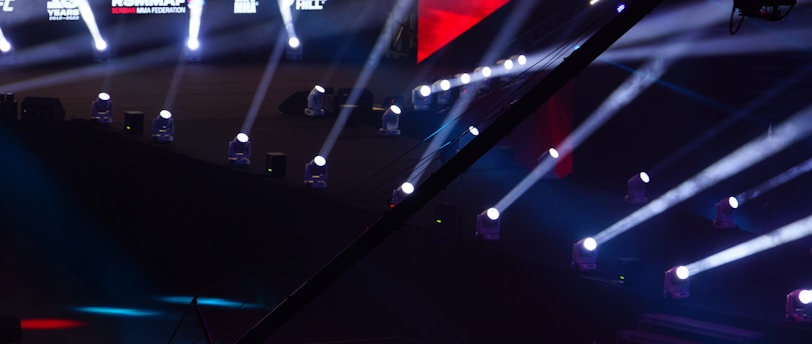 An array of bright stage lights beams across a dark arena, creating a dynamic atmosphere with a large screen displaying various logos and text. A camera crane is visible in the foreground, suggesting a professional event setup.