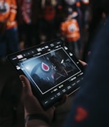 A person is holding a tablet that displays a video or image of a red and blue emblem on the screen. In the background, several people wearing dark and orange colored clothing, possibly sports jerseys, are partially visible. The scene is dimly lit, suggesting an indoor or evening setting.