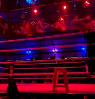 An empty boxing ring is illuminated by dramatic red and blue spotlights inside a dimly-lit arena. A small stool is placed in the center of the ring, surrounded by ropes. The arena is adorned with banners depicting action scenes, and a few silhouettes of spectators can be seen in the background.