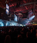 A large indoor event with a stage featuring bright screens and spotlights, attended by a substantial crowd. The venue has a high ceiling with industrial beams and red lighting accents. The atmosphere resembles a concert or a conference with a focus on visual effects.