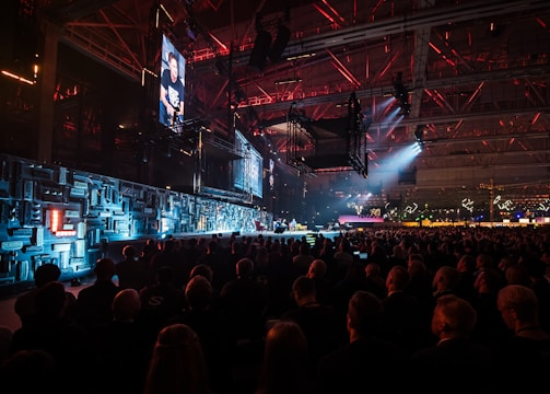 A large indoor event with a stage featuring bright screens and spotlights, attended by a substantial crowd. The venue has a high ceiling with industrial beams and red lighting accents. The atmosphere resembles a concert or a conference with a focus on visual effects.